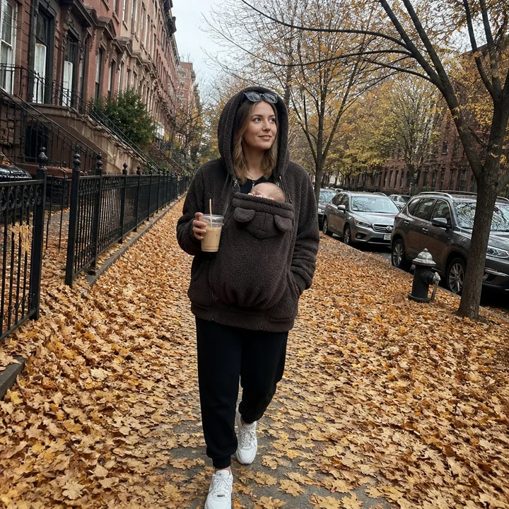 Femme en sweat marron foncé marchant dans une rue jonchée de feuilles, bébé dans le panneau, capuche relevée. 5