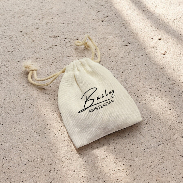Petite pochette en tissu écru à cordons, utilisée comme emballage du bijou, posée sur une surface sableuse éclairée par le soleil. 7