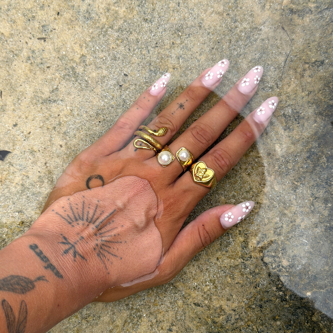 Main tatouée aux ongles fleuris, posée dans l'eau, portant la bague cœur dorée avec ange à côté d'une bague serpent et de bagues perlées. 4