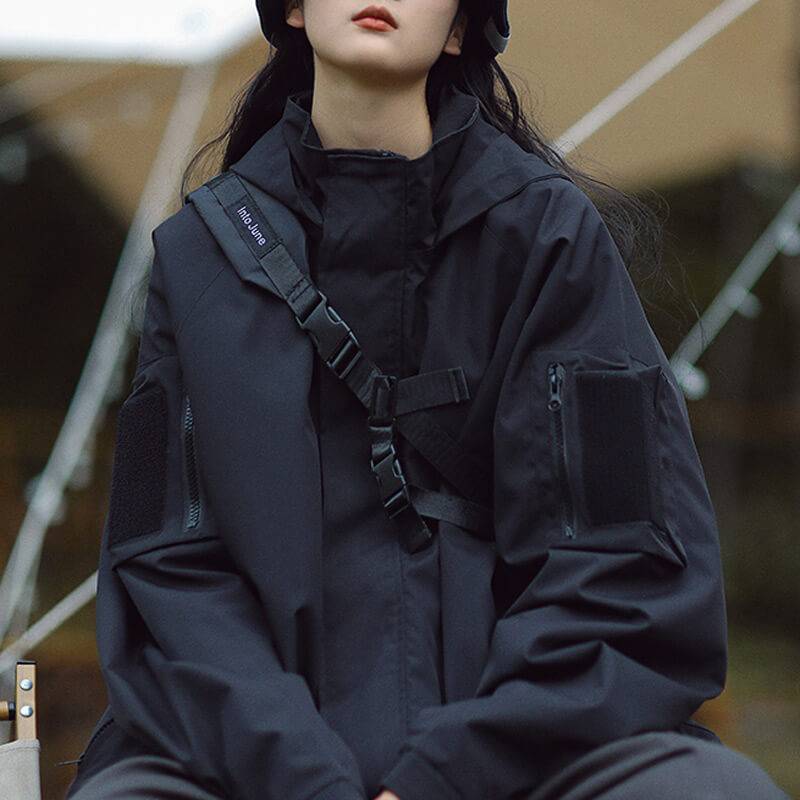 Femme assise en veste noire à capuche, avec une sangle à boucles en travers du buste et poches de manche à scratch. 5