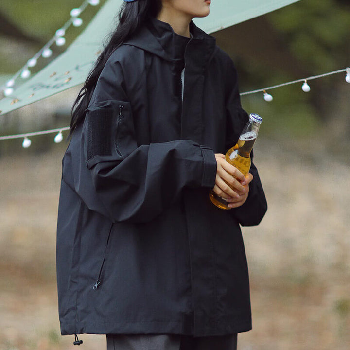 Femme en veste noire à capuche et poche zippée sur la manche, une bouteille à la main, guirlande lumineuse derrière. 1