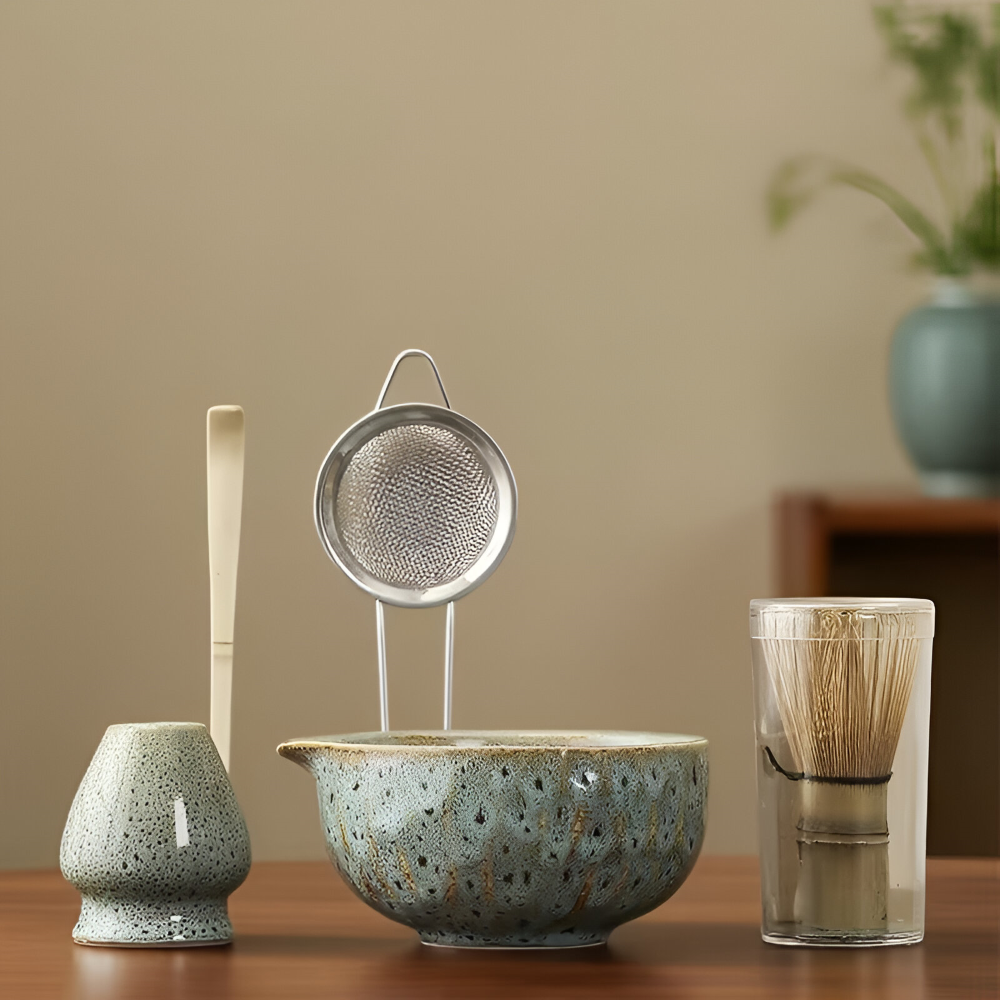 Pièces du set alignées sur une table en bois, bol, porte-fouet et passoire en versions vert-bleu moucheté, devant un mur beige. 1