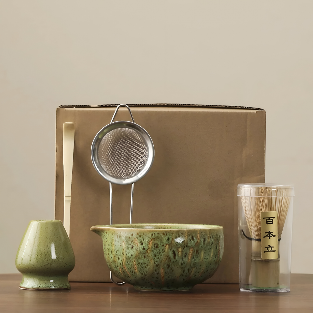 Pièces du set en céramique verte émaillée devant un carton d'emballage brun, avec passoire inox, cuillère et fouet en bambou. 4