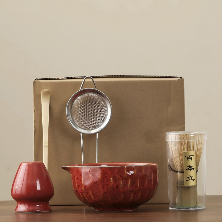 Pièces du set en céramique rouge vif brillant devant un carton d'emballage brun, avec passoire inox, cuillère et fouet en bambou. 13