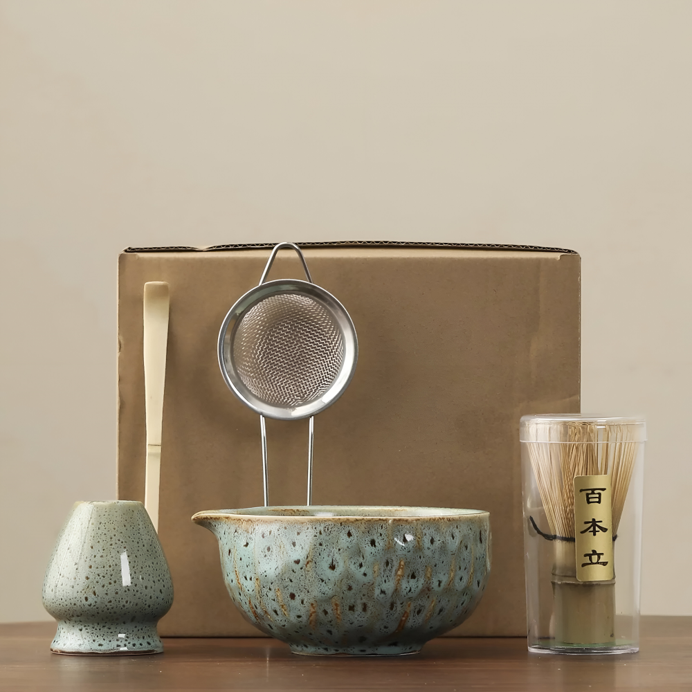 Pièces du set en céramique vert-bleu moucheté devant un carton d'emballage brun, avec passoire inox, cuillère et fouet en bambou. 14
