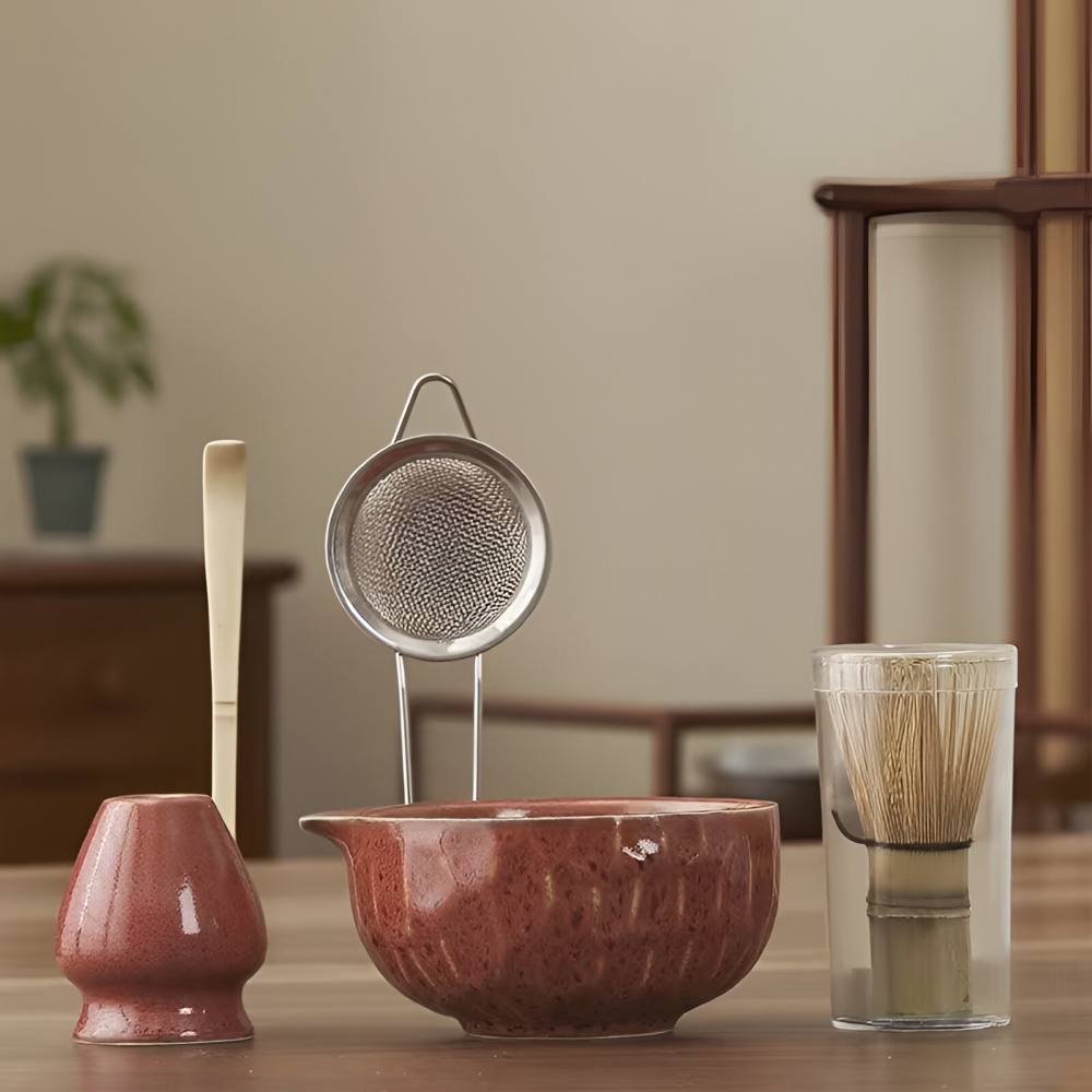 Pièces du set en céramique rouge brun brillant, avec passoire inox, cuillère en bambou et fouet protégé, dans un intérieur en bois. 2