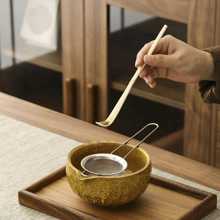 Main tenant la cuillère en bambou avec un peu de poudre de matcha au-dessus de la passoire posée dans le bol jaune. 9
