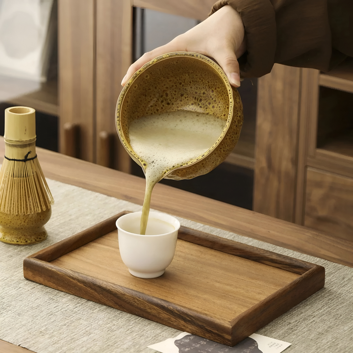 Matcha mousseux versé du bec du bol jaune dans une petite tasse blanche sur un plateau en bois, fouet à gauche. 10
