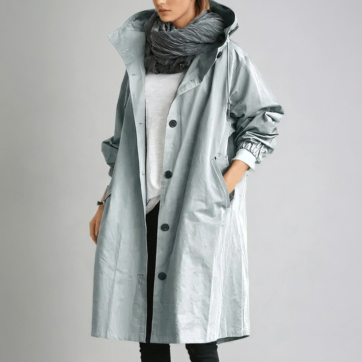 Imperméable long gris-bleu clair à capuche et boutons, porté avec une écharpe grise, une main dans la poche. 5