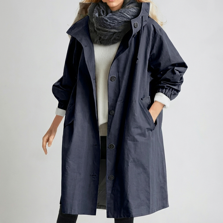 Imperméable long bleu marine à capuche, porté avec écharpe grise et pull blanc, une main dans la poche. 6