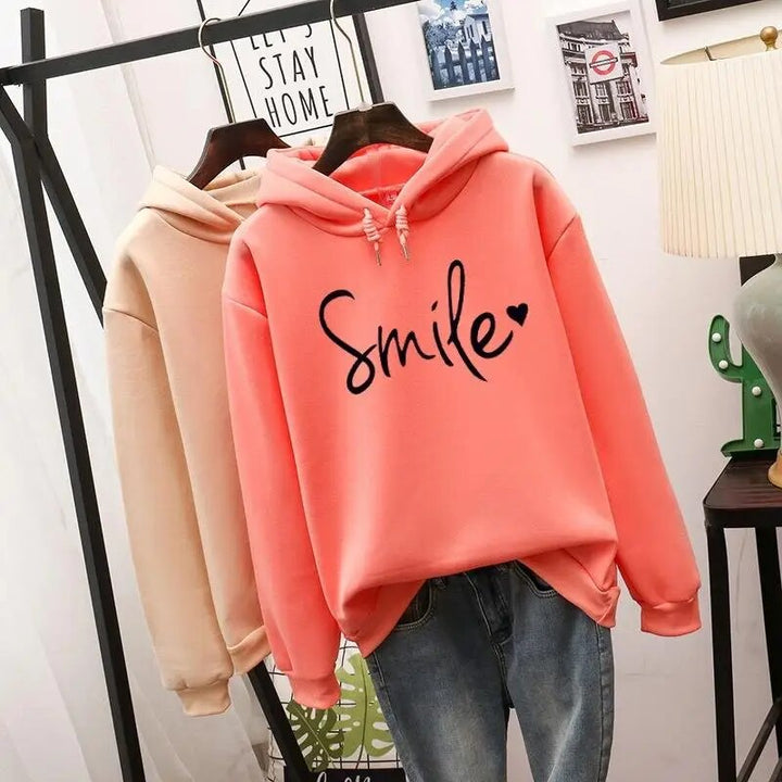 Sweat à capuche corail au premier plan avec motif noir et cœur, un second sweat beige derrière, sur portant. 3
