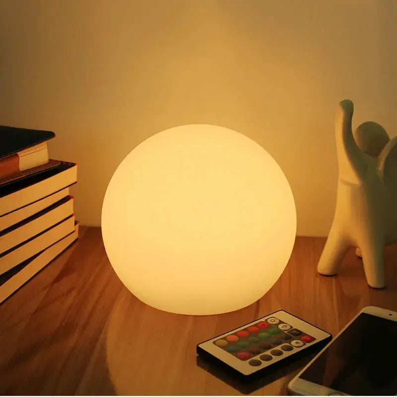 Orbe allumé en lumière chaude sur un bureau en bois, posé près d'une pile de livres, d'une télécommande colorée et d'un téléphone. 1