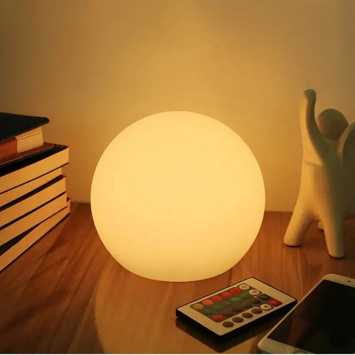 Orbe allumé en lumière chaude sur un bureau en bois, posé près d'une pile de livres, d'une télécommande colorée et d'un téléphone. 1