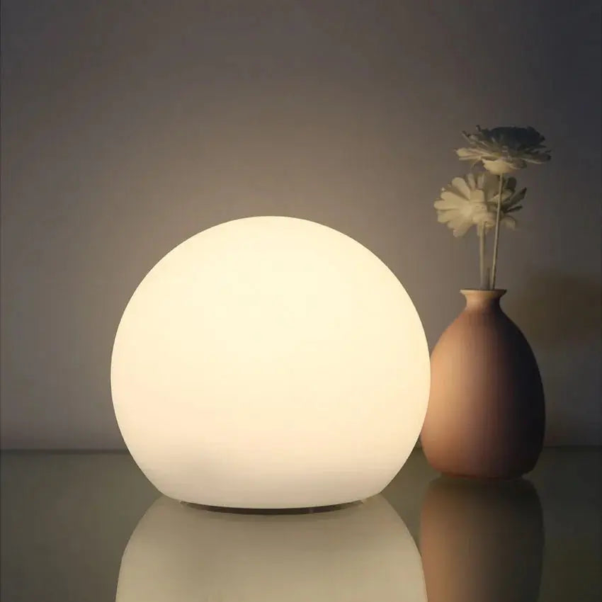 Orbe lumineux blanc chaud sur une surface brillante, à côté d'un petit vase beige contenant deux fleurs. 2