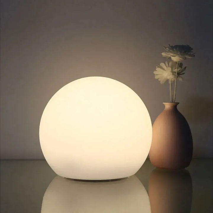 Orbe lumineux blanc chaud sur une surface brillante, à côté d'un petit vase beige contenant deux fleurs. 2