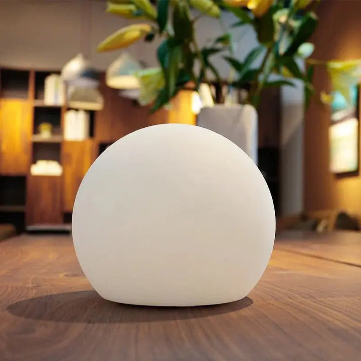 Orbe éteint, blanc mat, posé sur une table en bois dans un salon avec une plante et une bibliothèque en arrière-plan. 3