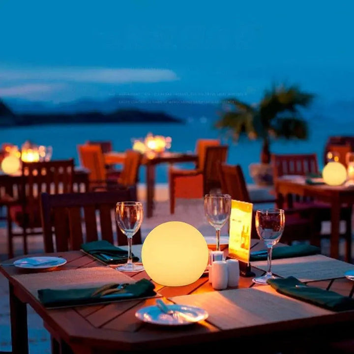Orbe jaune lumineux posé sur une table de restaurant en terrasse dressée avec verres et assiettes, au bord de la mer au crépuscule. 5