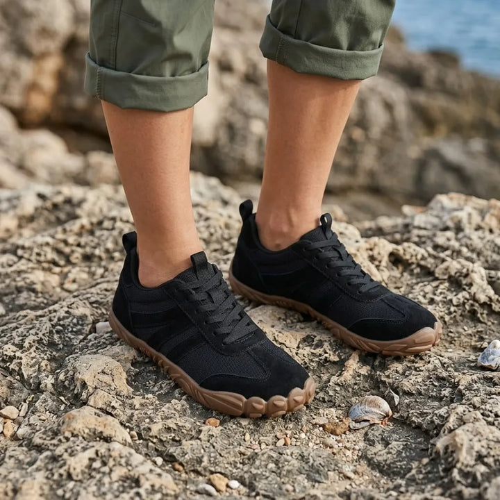 Pieds portant les chaussures noires, pantalon kaki retroussé, posés sur des rochers près de la mer avec quelques coquillages. 20
