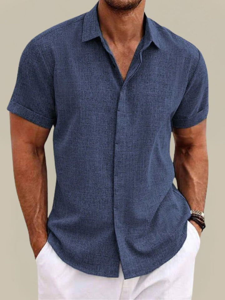 Homme portant la chemise bleu marine à manches courtes retroussées, boutonnée devant, avec un pantalon blanc, vue de face. 1