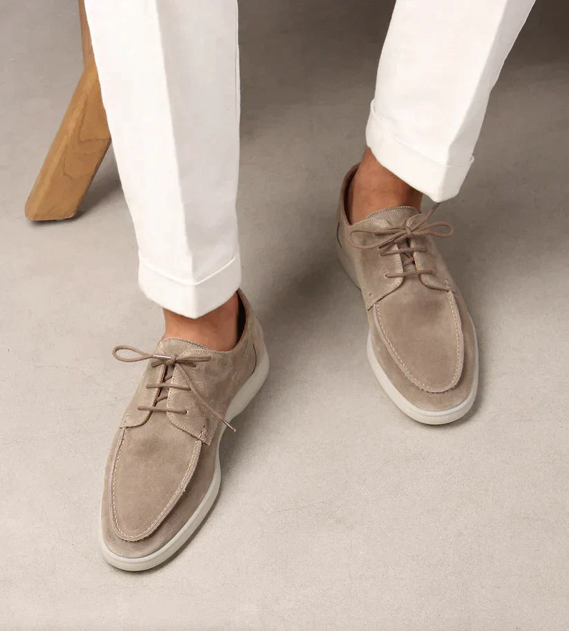 Vue de dessus de mocassins lacés en daim beige à semelle blanche, portés avec un pantalon blanc à revers, assis près d'un tabouret en bois. 2