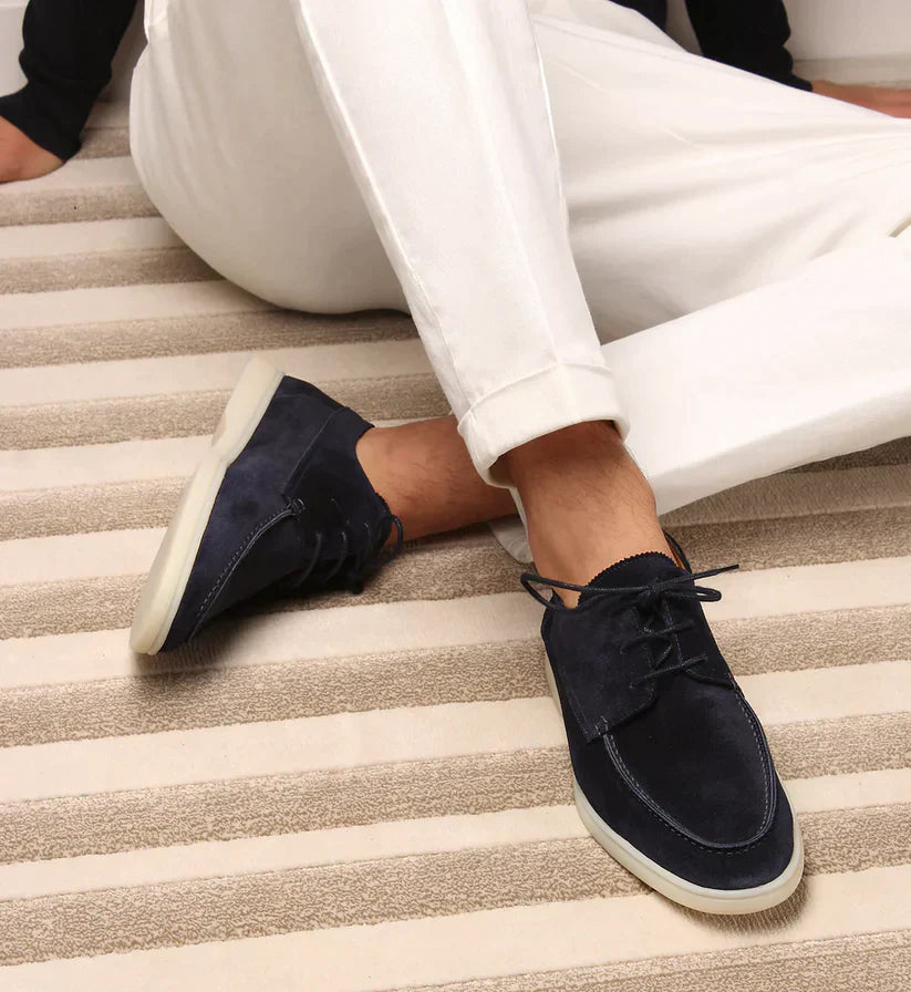 Homme assis sur un tapis rayé, chaussé de mocassins lacés en daim bleu marine à semelle blanche, avec un pantalon blanc. 1