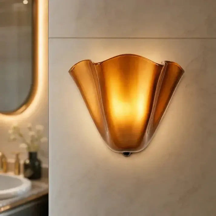 Applique ambrée allumée sur un mur de salle de bain en pierre claire, avec un miroir lumineux flou à gauche. 10