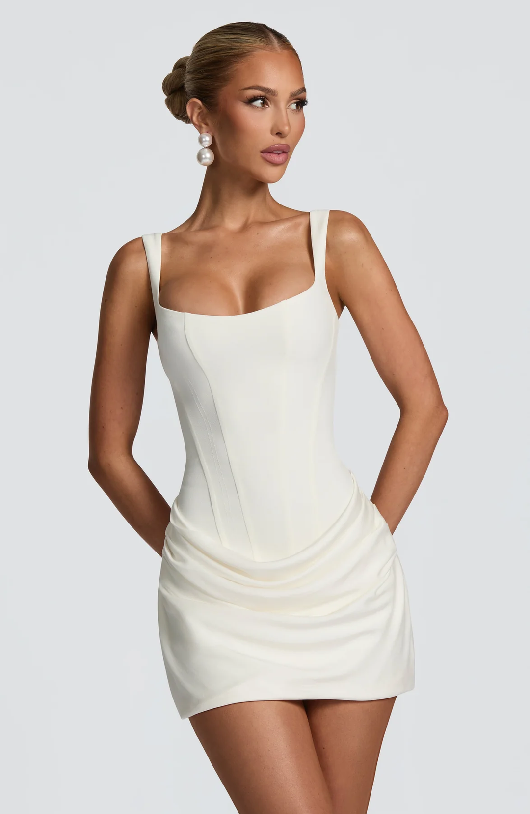 Gros plan de face sur une mannequin portant la robe crème à bustier et jupe drapée, mains dans le dos, boucles d'oreilles perles. 1