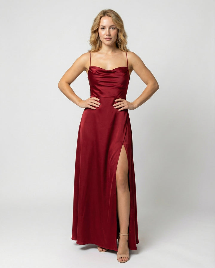 Femme blonde portée de face, mains sur les hanches, en robe longue satinée rouge bordeaux avec décolleté drapé et fente haute. 1