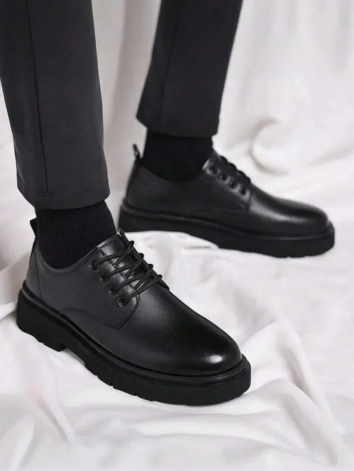 Homme debout portant les derbies noires avec pantalon gris et chaussettes noires, sur fond de tissu blanc 1