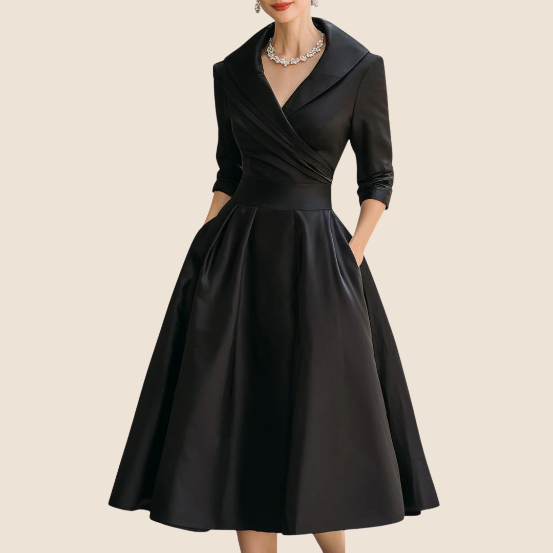 Robe noire portée de face par une femme, col châle croisé, manches trois-quarts, mains dans les poches, collier de strass. 1