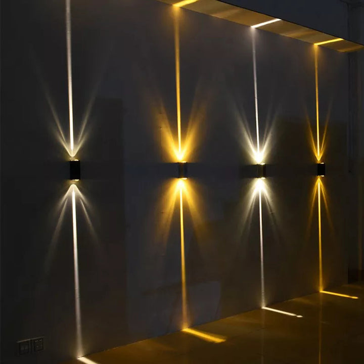 Quatre appliques noires fixées sur un mur sombre projetant des faisceaux lumineux fins, blancs et jaunes, vers le haut et le 1