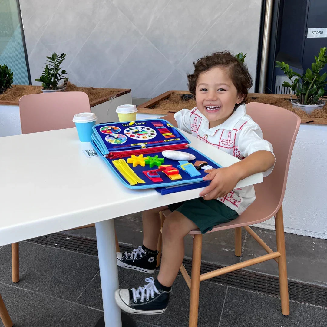 Garçon rieur assis à une table blanche en terrasse, le livre d'activités bleu ouvert devant lui avec horloge, engrenages et fermeture éclair jaune. 7
