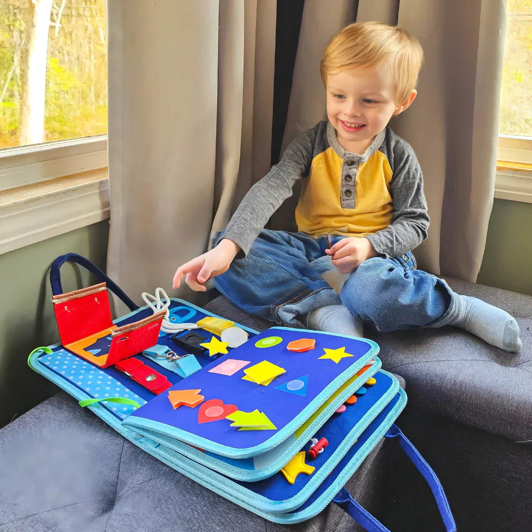 Petit garçon blond souriant assis près d'une fenêtre, qui joue avec le livre d'activités bleu ouvert posé devant lui, formes en feutrine visibles. 3
