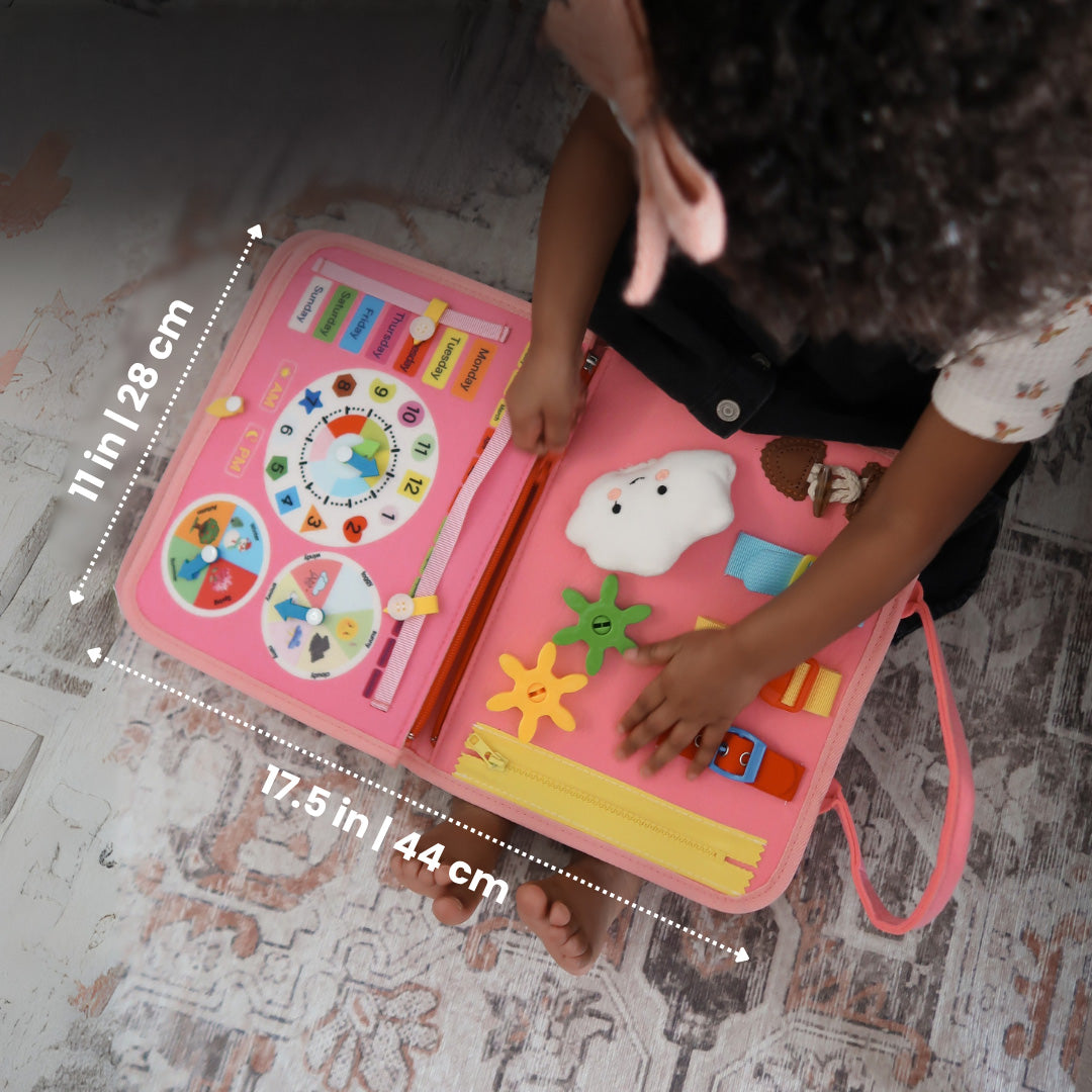 Vue du dessus du livre d'activités rose ouvert, tenu par une enfant, avec ses dimensions indiquées en centimètres et en pouces, horloge et nuage en peluche. 8
