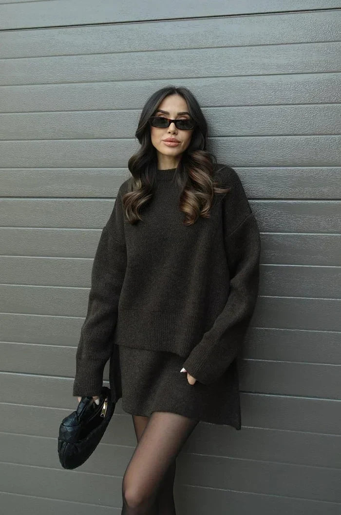 Ensemble en maille marron foncé porté de côté, mannequin adossée au mur, une main dans la poche, pochette noire tressée. 13