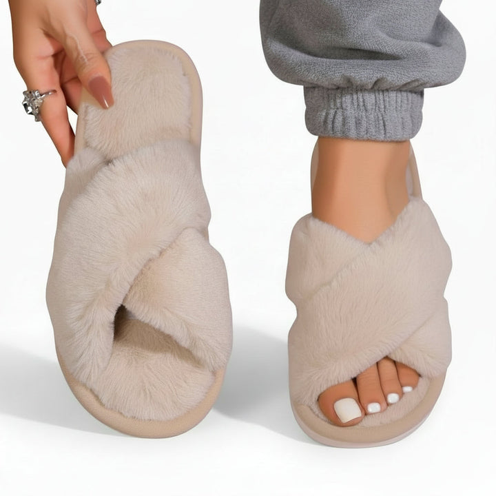 Chaussons beige clair à brides croisées en peluche, un porté avec un pantalon de jogging gris et l'autre tenu à la main. 7
