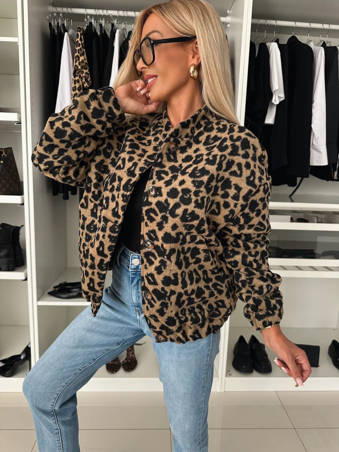 Femme blonde portant la veste boutonnée à imprimé léopard sur un jean, vue de trois quarts, main relevée vers le visage, dans un dressing. 1