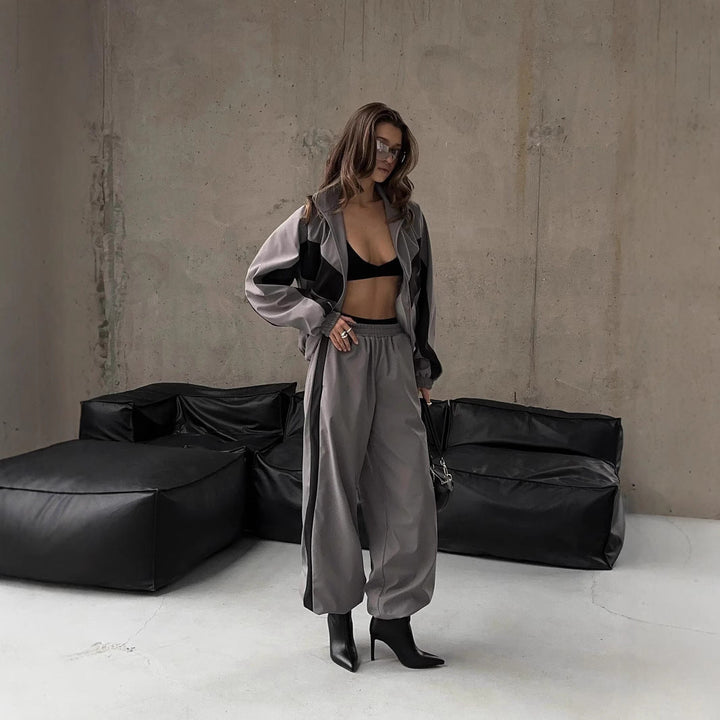Modèle debout devant des poufs noirs, ensemble gris à empiècements noirs, veste ouverte et pantalon large resserré aux chevilles. 7