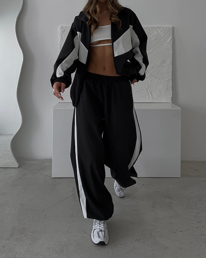 Femme en veste zippée noire à empiècements blancs et pantalon large noir à bande latérale blanche, avec baskets blanches. 21