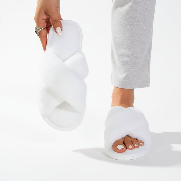 Chaussons blancs à brides croisées en peluche, un porté avec un pantalon gris clair et l'autre tenu à la main. 6