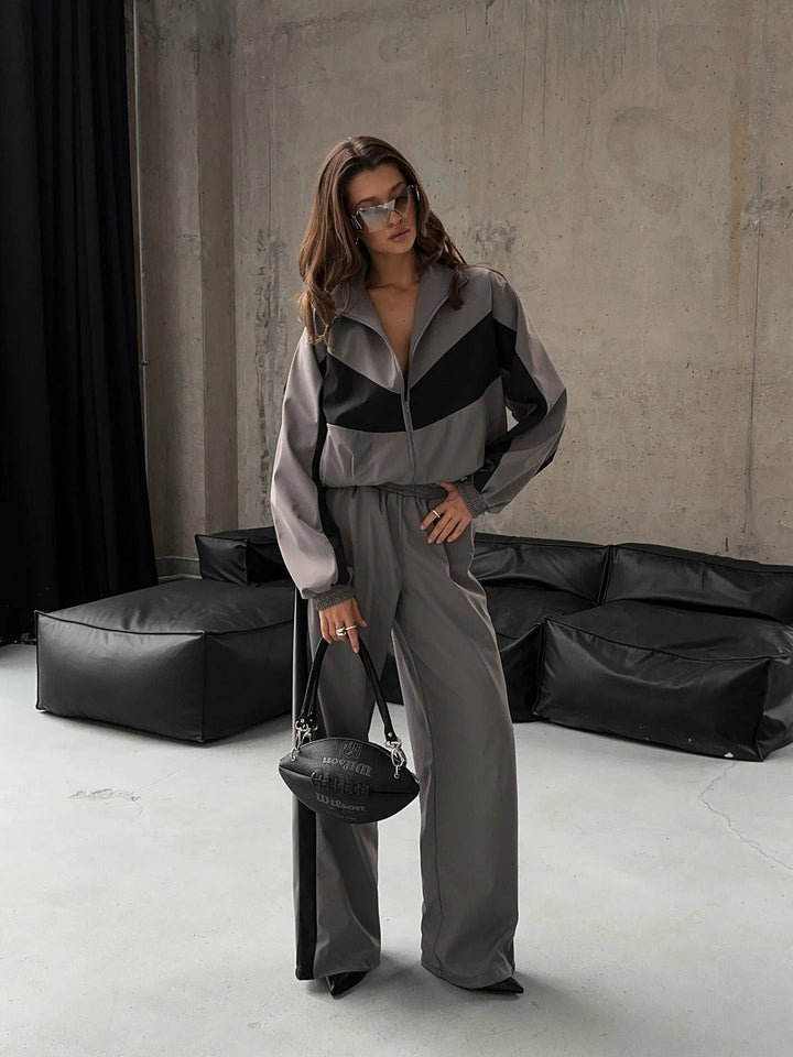 Femme debout en ensemble gris à empiècements noirs, veste zippée et pantalon large, lunettes de soleil et sac noir à la main. 23