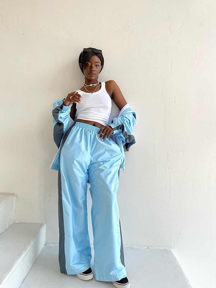 Modèle adossée à un mur blanc en pantalon large bleu clair à bande grise, veste assortie portée ouverte sur les épaules. 14