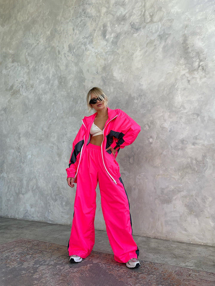 Modèle debout devant un mur texturé, ensemble rose fluo à bandes gris foncé, veste zippée ouverte et pantalon large. 18