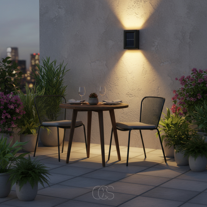 Applique solaire allumée sur le mur en crépi d'une terrasse, éclairant vers le haut et le bas une table de bistrot dressée pour deux. 1