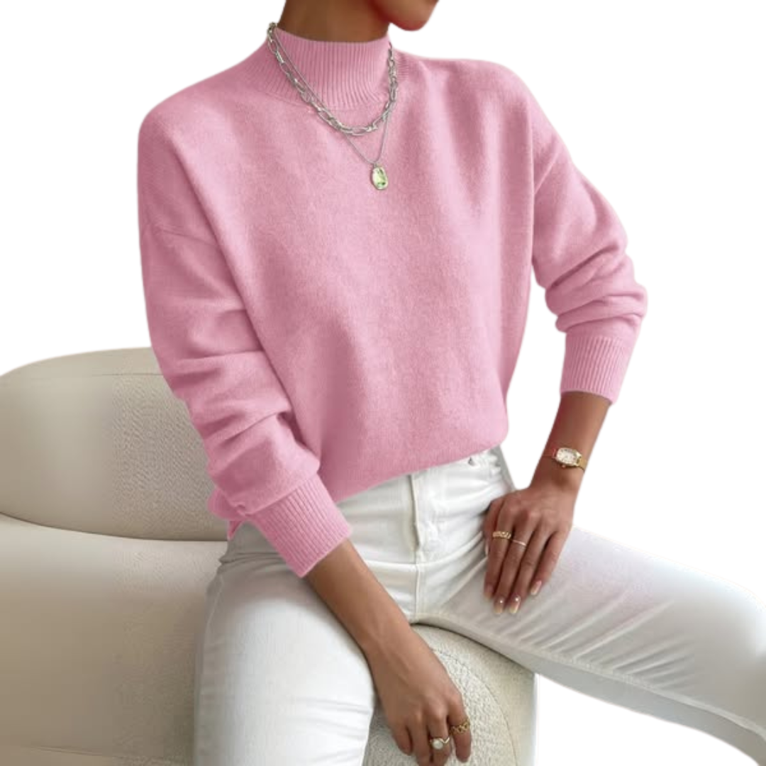 Pull rose clair à col montant porté par une femme assise, avec pantalon blanc et collier argenté, fond blanc. 1