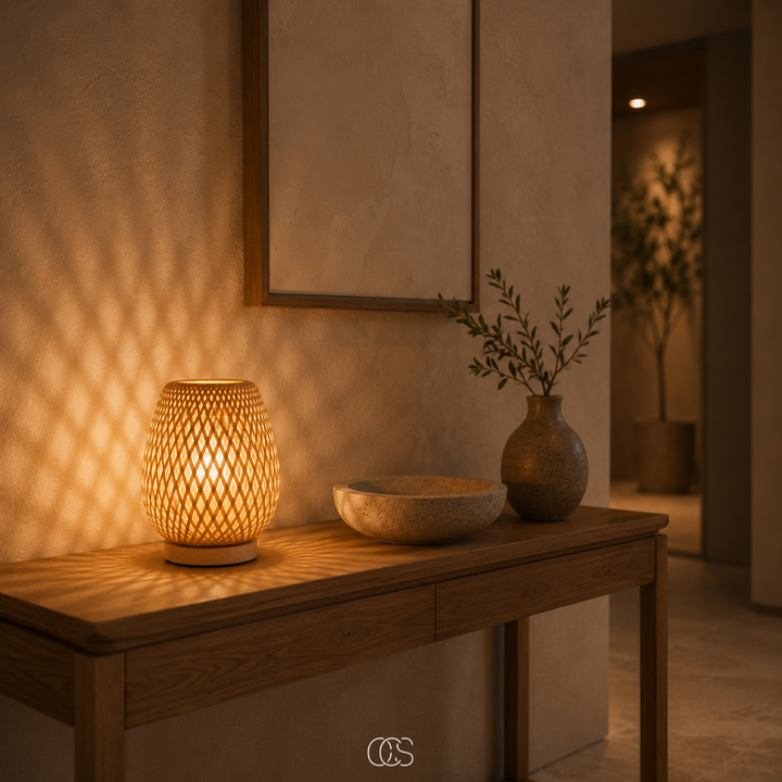 Lampe en bambou tressé allumée sur une console en bois, projetant des motifs lumineux sur le mur, à côté d'une coupe et d'un vase. 2