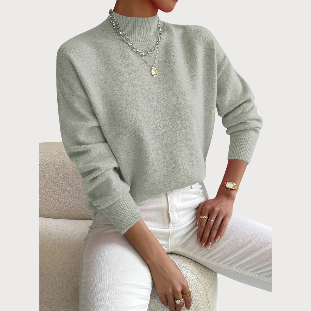 Pull vert sauge à col montant et poignets côtelés, porté avec un pantalon blanc, assise sur un fauteuil bouclé crème. 2
