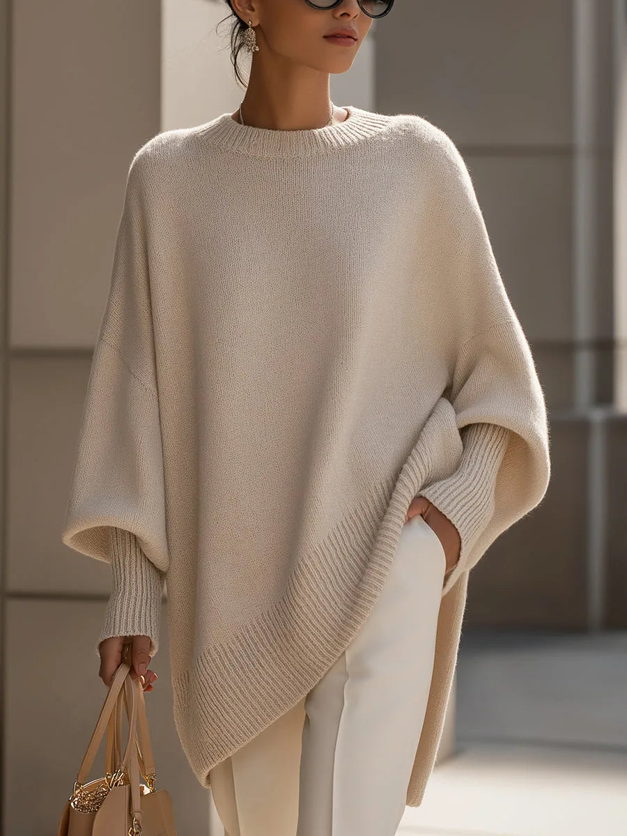 Pull oversize beige en maille porté par une femme avec un pantalon blanc, vue de face, ourlet côtelé asymétrique et manches amples. 1