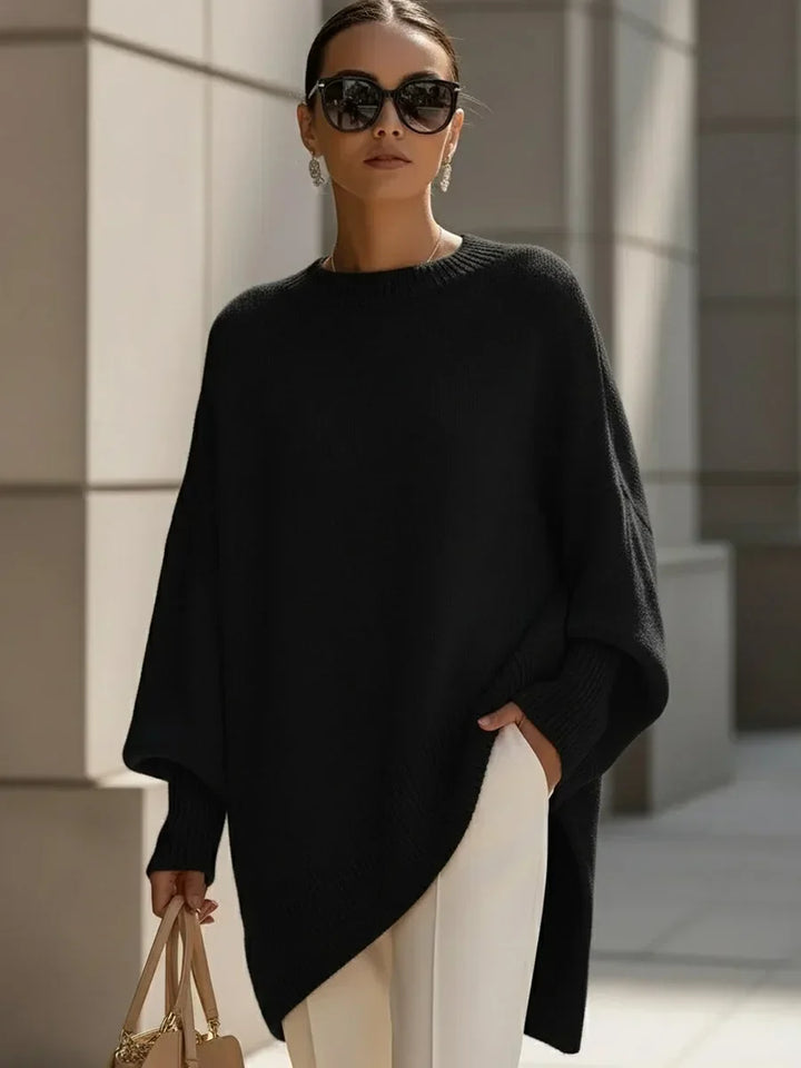 Pull noir oversize à ourlet asymétrique porté avec un pantalon blanc, lunettes de soleil et sac beige, vue de face. 6