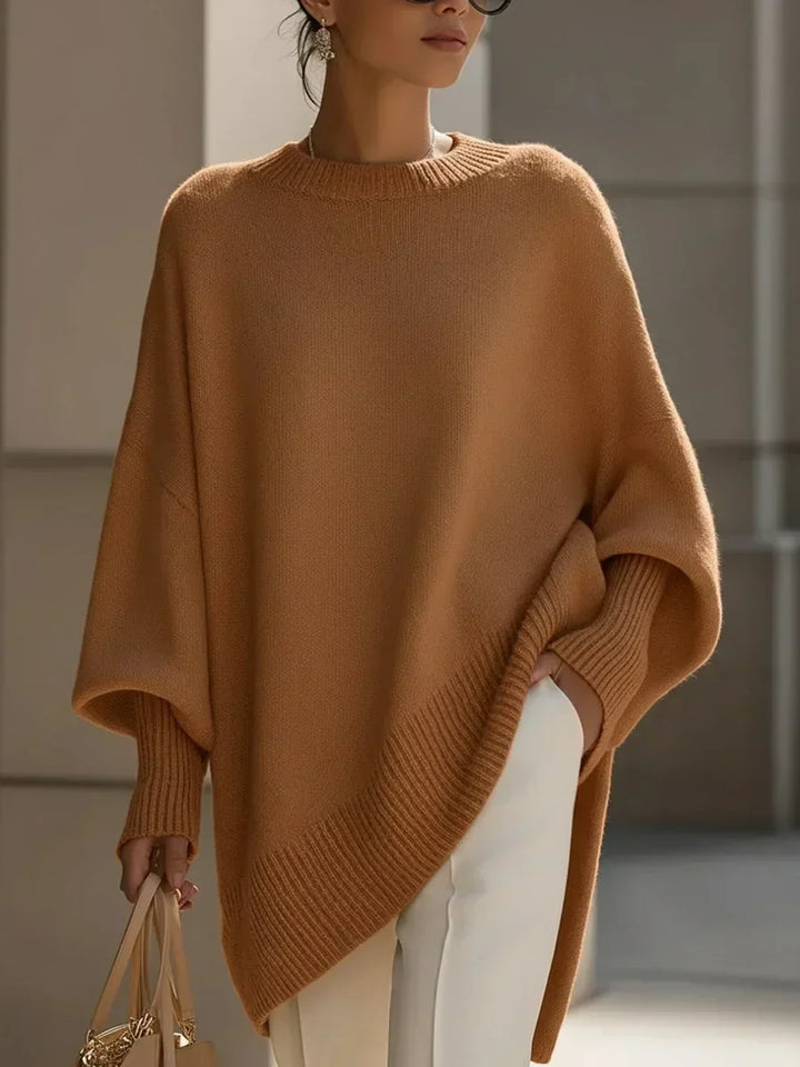 Pull camel oversize à manches amples et ourlet asymétrique côtelé, porté avec un pantalon blanc, main dans la poche. 12